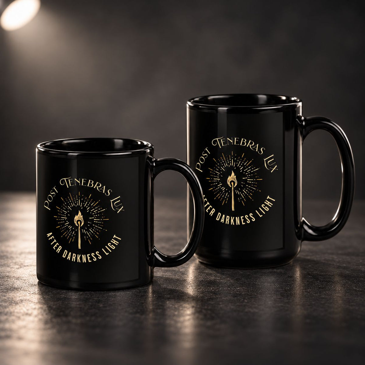 Two black ceramic mugs with gold printed graphics reading 'POST TENEBRAS LUX' and 'AFTER DARKNESS LIGHT' around a central flame motif, shown on a dark tabletop against a dim background.