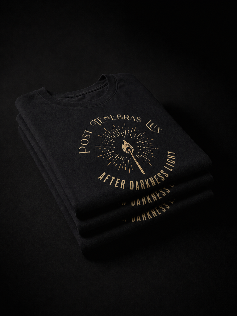 A stack of folded black T-shirts on a black background, featuring a gold graphic print with a central eye motif and the text 'Post Tenebras Lus' and 'AFTER DARKNESS LIGHT'.