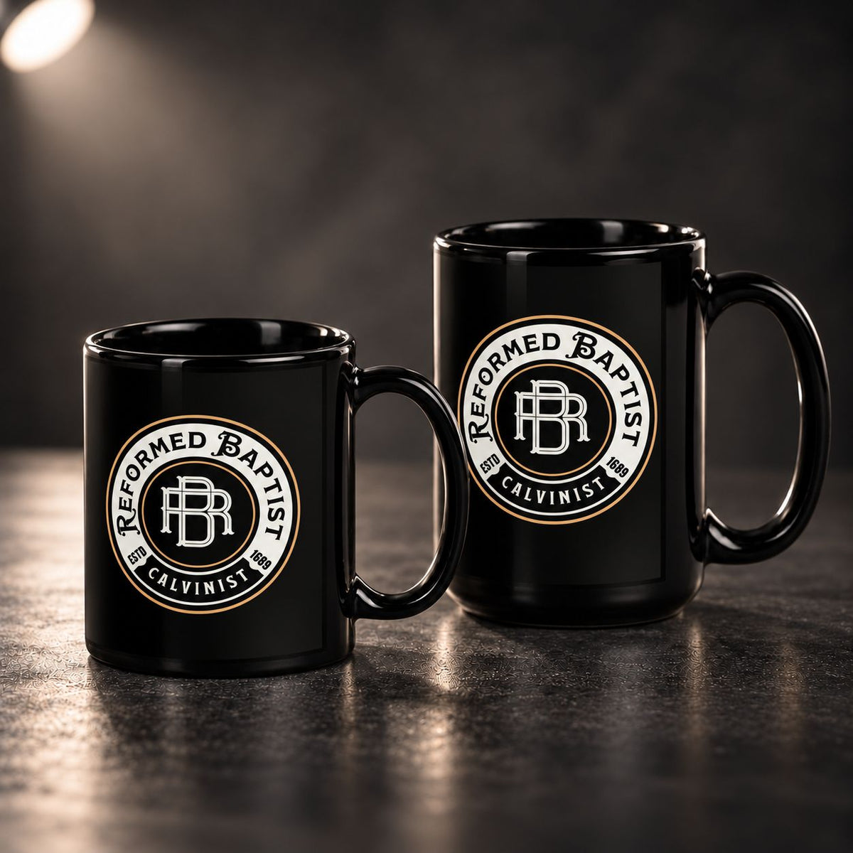 Two black ceramic mugs with a circular 'Reformed Baptist Calvinist' logo, displayed on a dark reflective surface against a dark background.